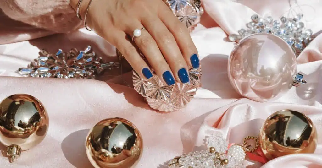 Stylish hand showcasing blue nails resting on pink satin surrounded by opulent ornaments and jewelry.