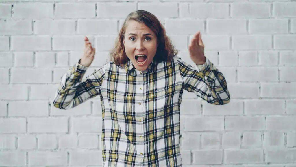 Shocked woman with raised hands in plaid shirt, standing indoors against a white brick wall.